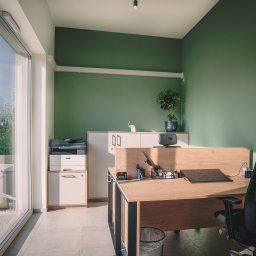 P.H.U. Konrad Kiełkowski - Nowoczesne biuro po malowaniu ścian na zielono, z dużym oknem, biurkiem z laptopem i ergonomcznym krzesłem. Widoczny fragment klimatyzacji za oknem i roślina na szafce.