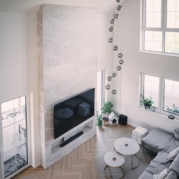 P.H.U. Konrad Kiełkowski - Salon z parkietem ułożonym w jodełkę, ściana TV wykończona marmurem, wisząca lampa z kul, okna z roślinami, szara sofa. Widok z góry na wykończone wnętrze.
