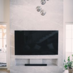 P.H.U. Konrad Kiełkowski - Elegancka ściana TV z marmurowym wykończeniem i wnęką na soundbar. Nad telewizorem wiszą cztery chromowane kule-lampy. Parkiet ułożony w jodełkę.