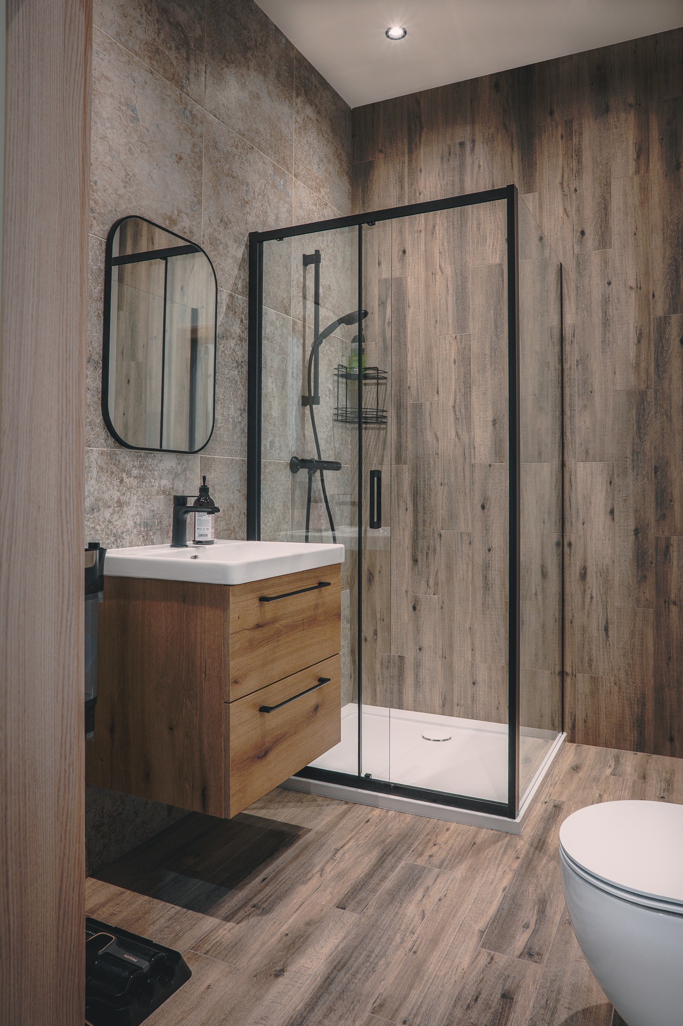 Nowoczesna łazienka z kabiną prysznicową w czarnej ramie, drewnianą szafką pod umywalką i lustrem. Płytki imitujące drewno na podłodze i ścianach.