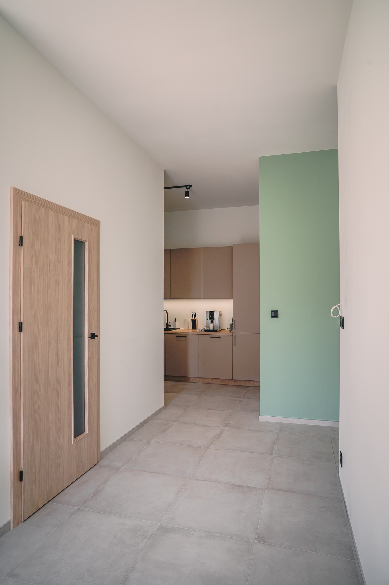 Nowoczesne wnętrze: jasna dębowa stolarka drzwiowa z przeszkleniem, szare płytki podłogowe, ściana w kolorze pistacjowym i zabudowa kuchenna w tle.