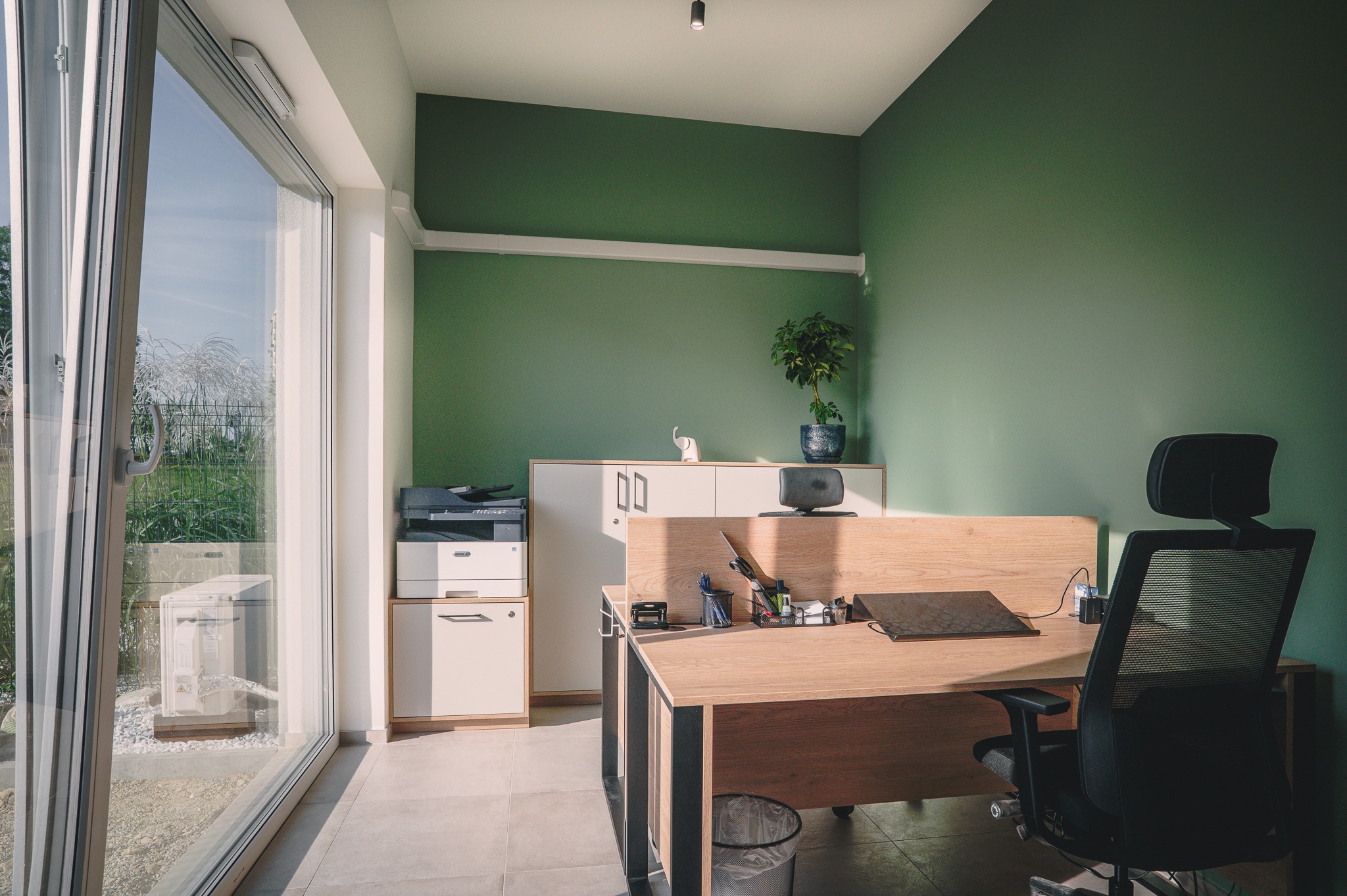 Nowoczesne biuro po malowaniu ścian na zielono, z dużym oknem, biurkiem z laptopem i ergonomcznym krzesłem. Widoczny fragment klimatyzacji za oknem i roślina na szafce.