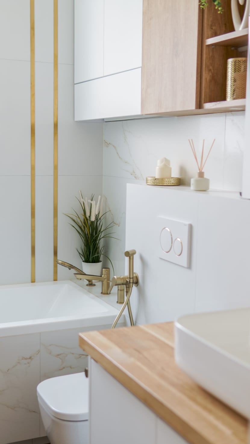 Elegancka łazienka z wanną obłożoną marmurem, złotą armaturą i dekoracyjnymi złotymi paskami na białych płytkach. Widoczna toaleta i drewniany blat umywalki.