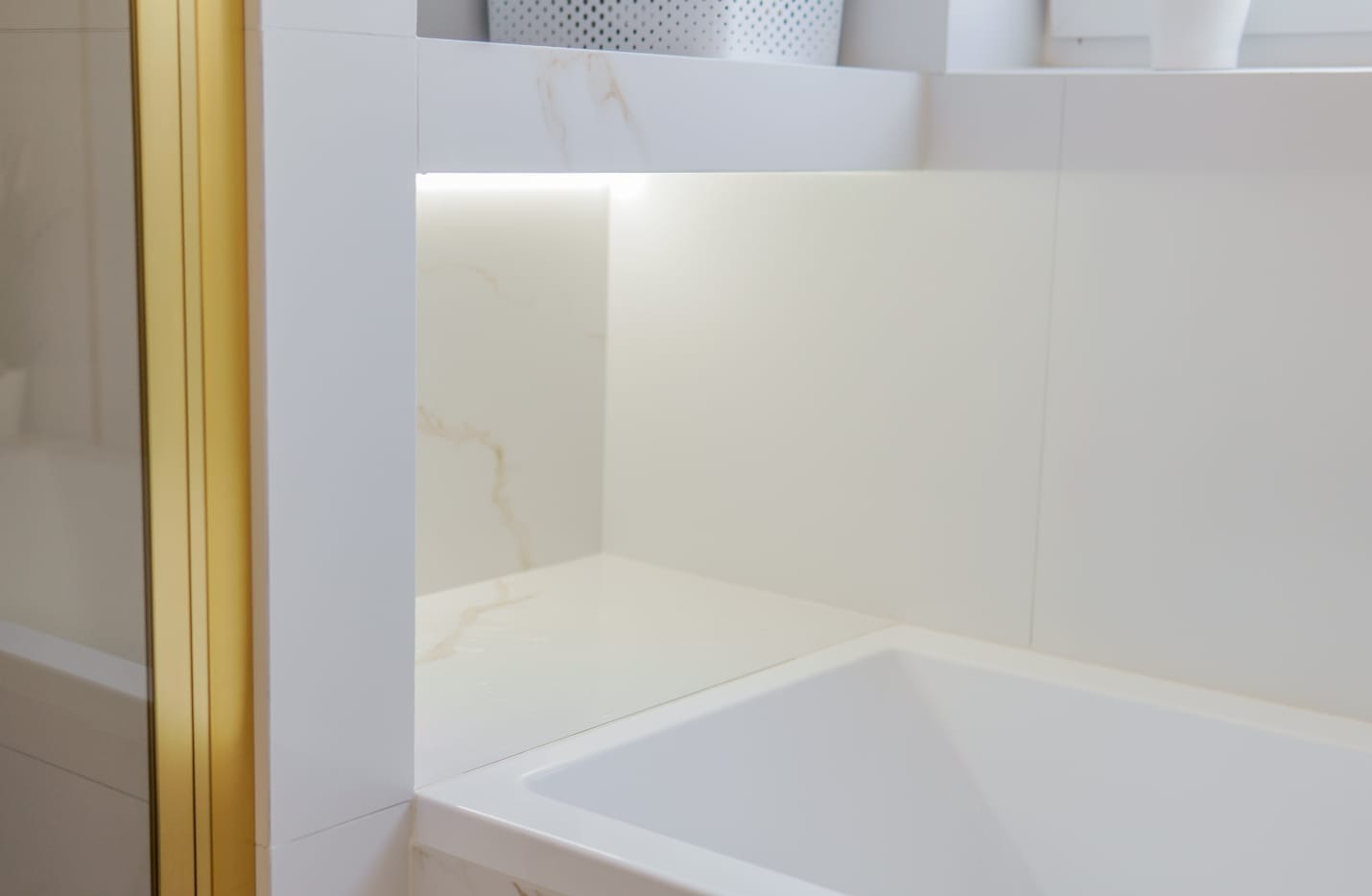 Nowoczesna łazienka z białą wanną, złotymi detalami kabiny i wnęką z podświetleniem LED. Minimalistyczny design z płytkami imitującymi marmur.