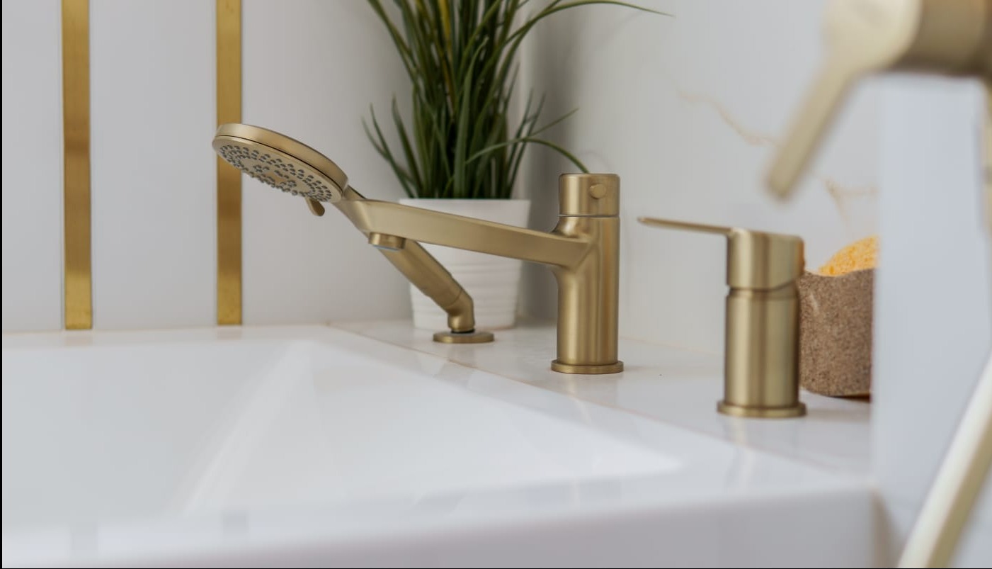 Złota bateria wannowa z deszczownicą na białej wannie, na tle białych płytek z złotymi paskami i rośliną w doniczce. Nowoczesny design łazienki.