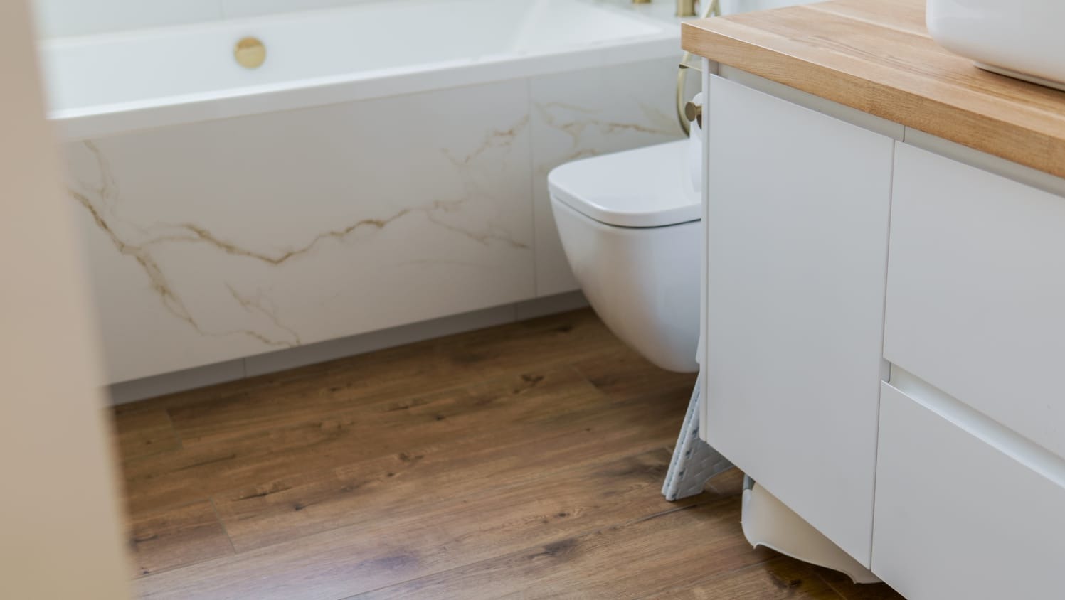 Nowoczesna łazienka: drewnopodobne panele, biała armatura i szafka z blatem drewnianym. Minimalistyczny design z marmurowym akcentem na obudowie wanny.