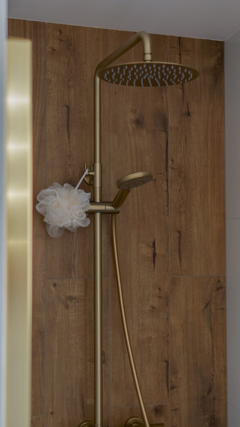 Złota deszczownica z słuchawką prysznicową na tle drewnopodobnych płytek. Na baterii zawieszona myjka. Nowoczesny design.