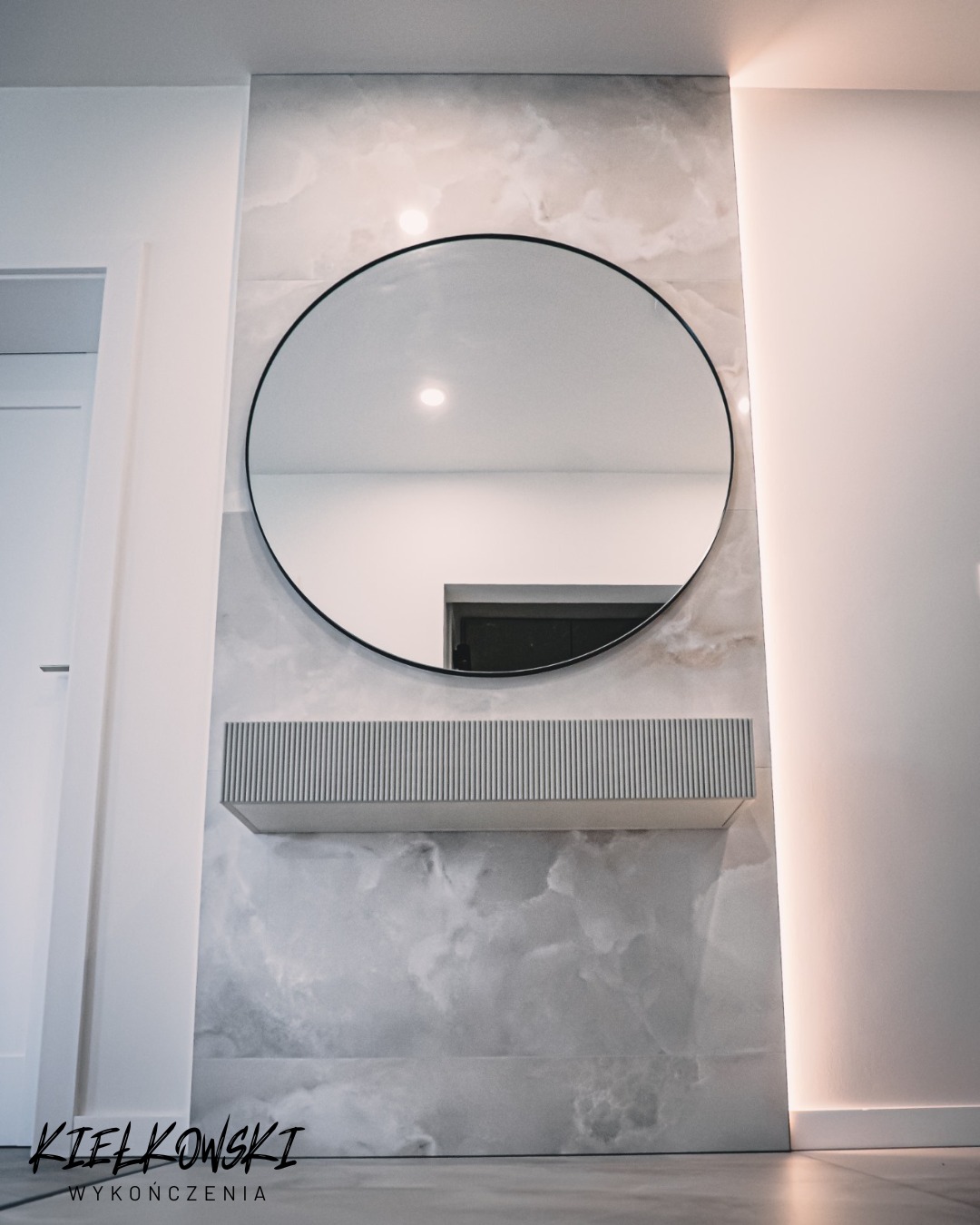 Nowoczesny przedpokój z okrągłym lustrem i półką na tle ściany imitującej marmur. Minimalistyczny design, subtelne oświetlenie LED. Wykończenia w bieli i szarości.