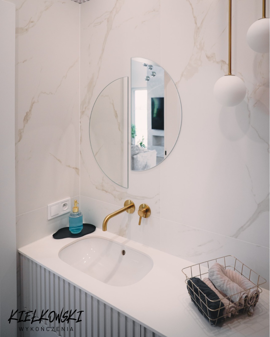 Elegancka łazienka z umywalką, złotą armaturą i okrągłym lustrem podzielonym na pół. Marmurowe płytki na ścianie, minimalistyczny styl.