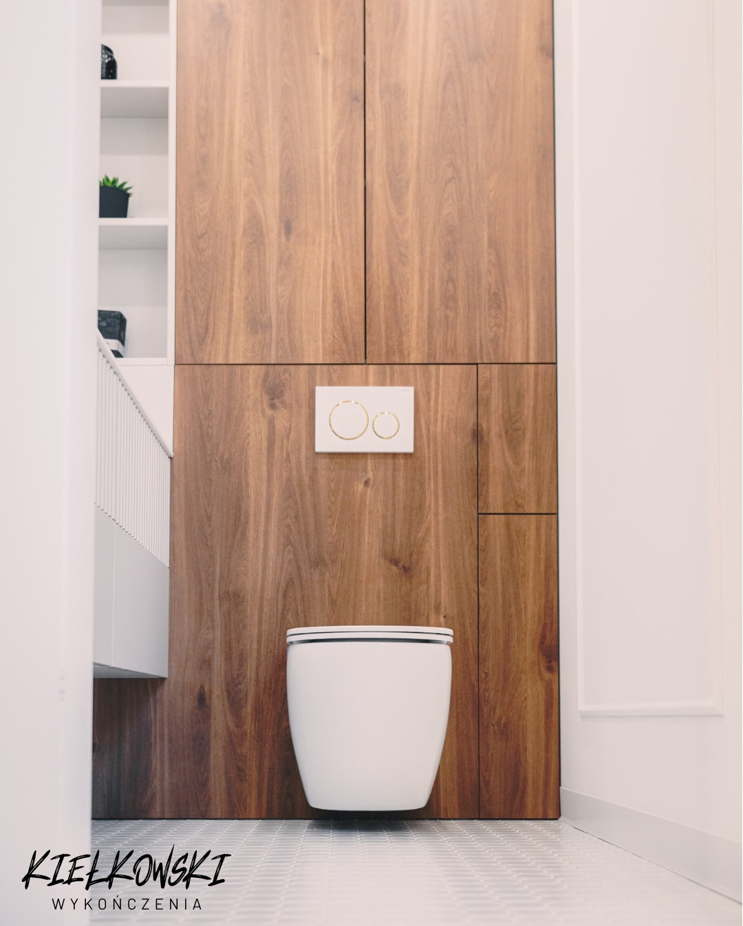 Nowoczesna toaleta podwieszana na tle drewnianej zabudowy, z widocznym przyciskiem spłukującym i fragmentem białej szafki. Minimalistyczne wnętrze łazienki.