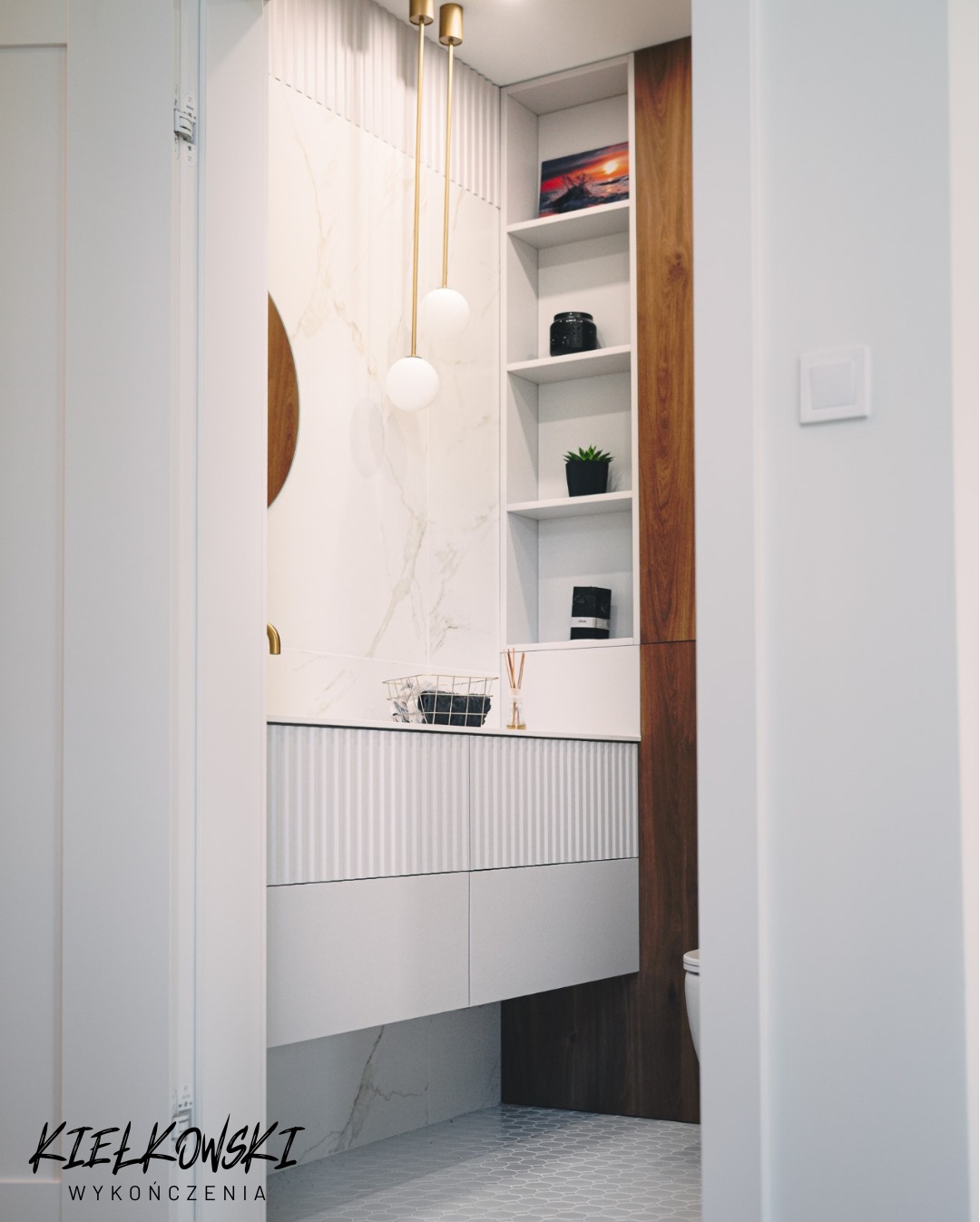 Nowoczesna łazienka z marmurową ścianą, wiszącą szafką z ryflowanym frontem i złotymi lampami. Detale obejmują drewniany regał i heksagonalną podłogę.