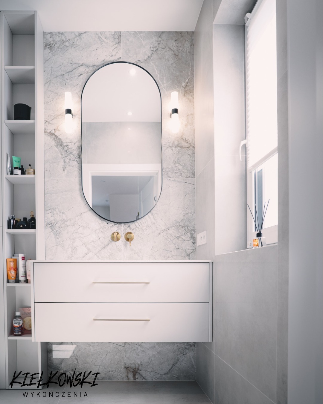 Nowoczesna łazienka z owalnym lustrem, białymi szafkami pod umywalką i marmurowymi płytkami. Widoczne oświetlenie punktowe i okno z roletą.