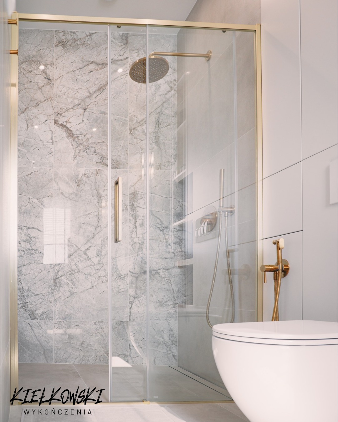 Nowoczesna kabina prysznicowa z marmurowymi płytkami i złotymi detalami, widok na toaletę i bidetkę w jasnej łazience.