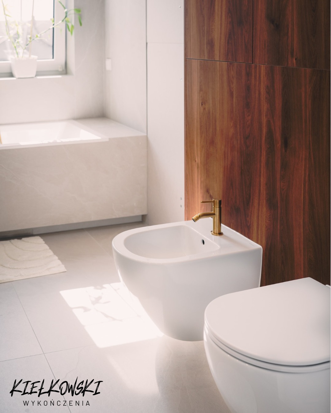 Nowoczesna łazienka z białym bidetem i toaletą, złota armatura, drewniana zabudowa, jasne płytki podłogowe i subtelne oświetlenie okienne.