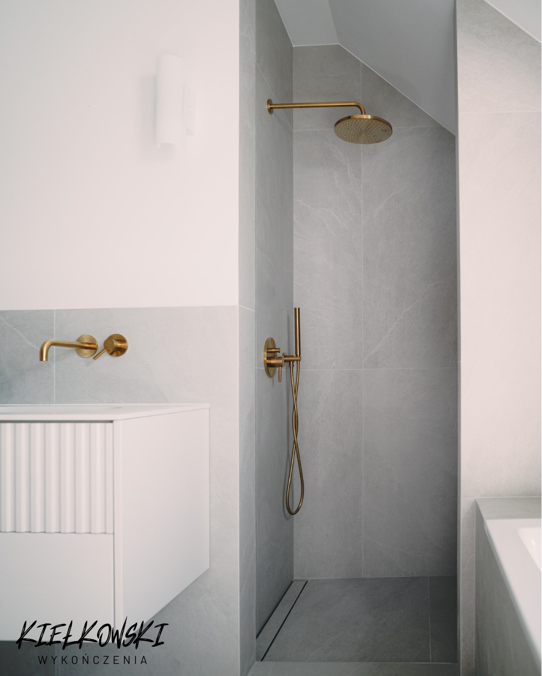 Szara łazienka z kabiną typu walk-in, złota armatura, umywalka z szafką. Uwagę przykuwa odpływ liniowy w podłodze kabiny i minimalistyczna lampa ścienna.