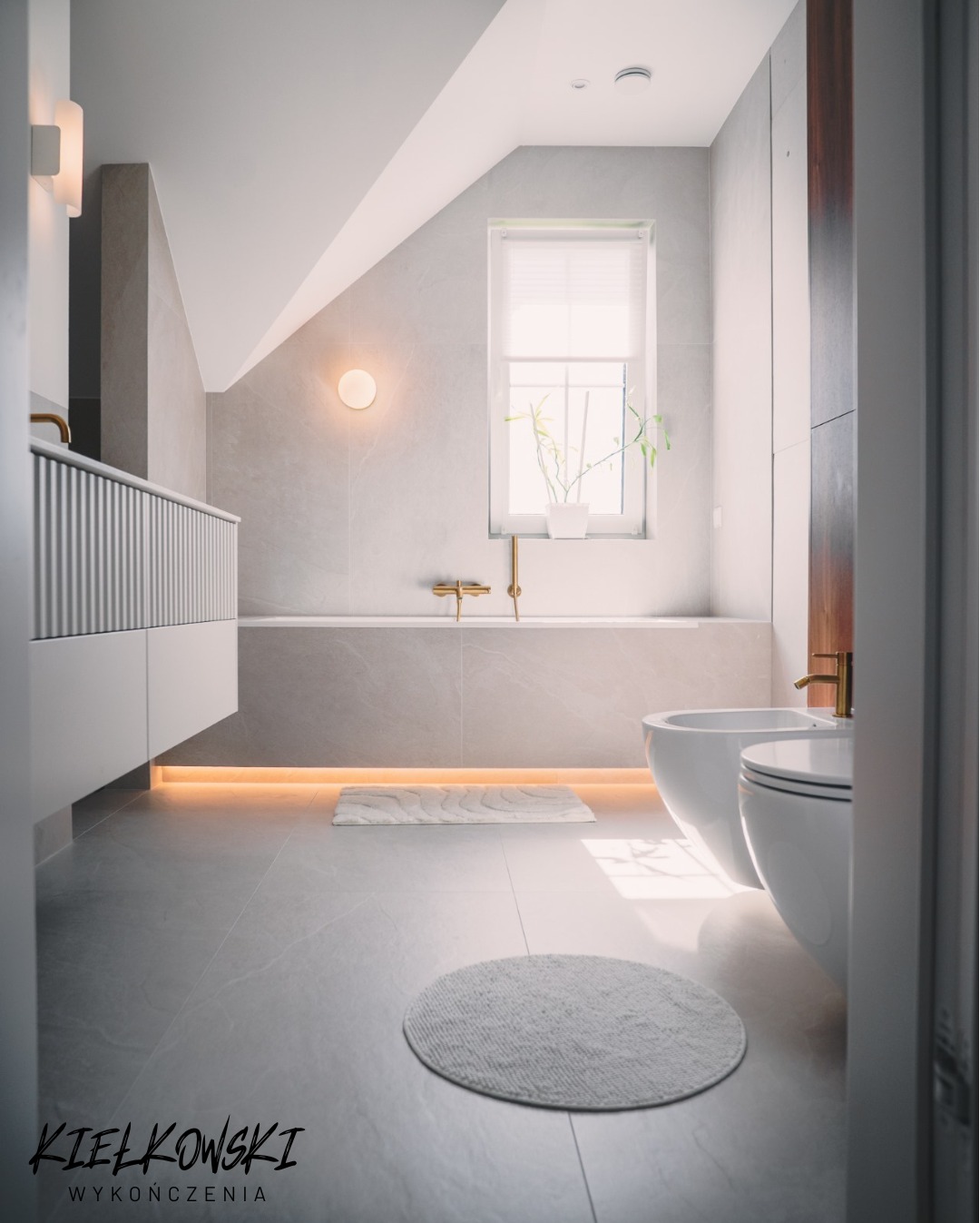 Nowoczesna łazienka na poddaszu z szarą płytką, wanną, bidetem i WC. Złota armatura i podświetlenie LED dodają elegancji. Widoczny dywanik i roślina w oknie.
