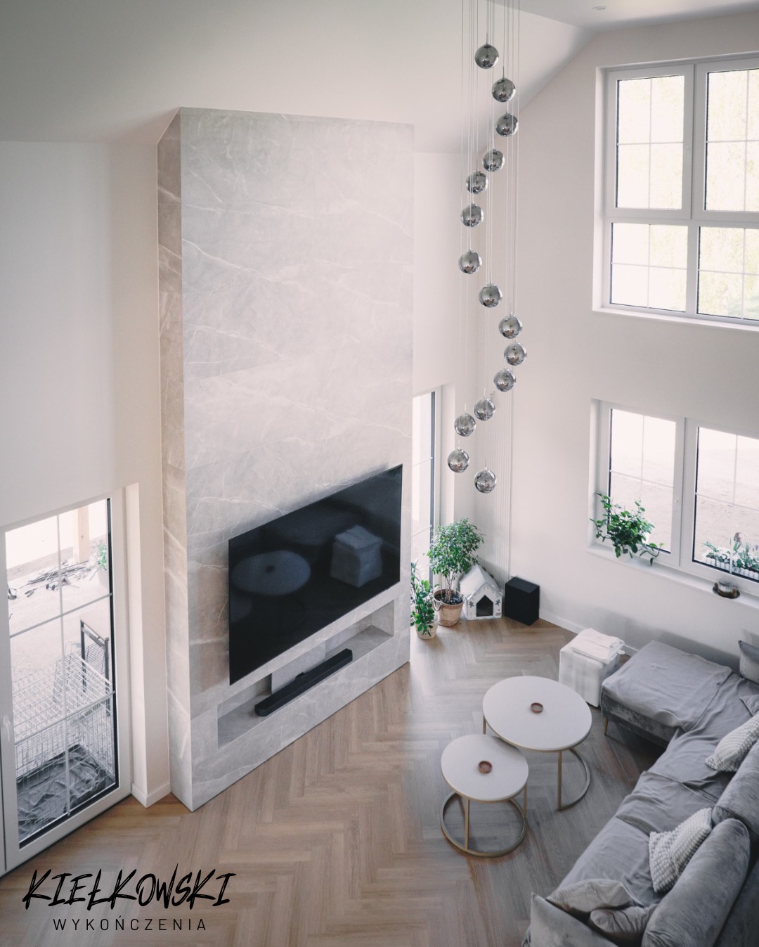 Salon z parkietem ułożonym w jodełkę, ściana TV wykończona marmurem, wisząca lampa z kul, okna z roślinami, szara sofa. Widok z góry na wykończone wnętrze.