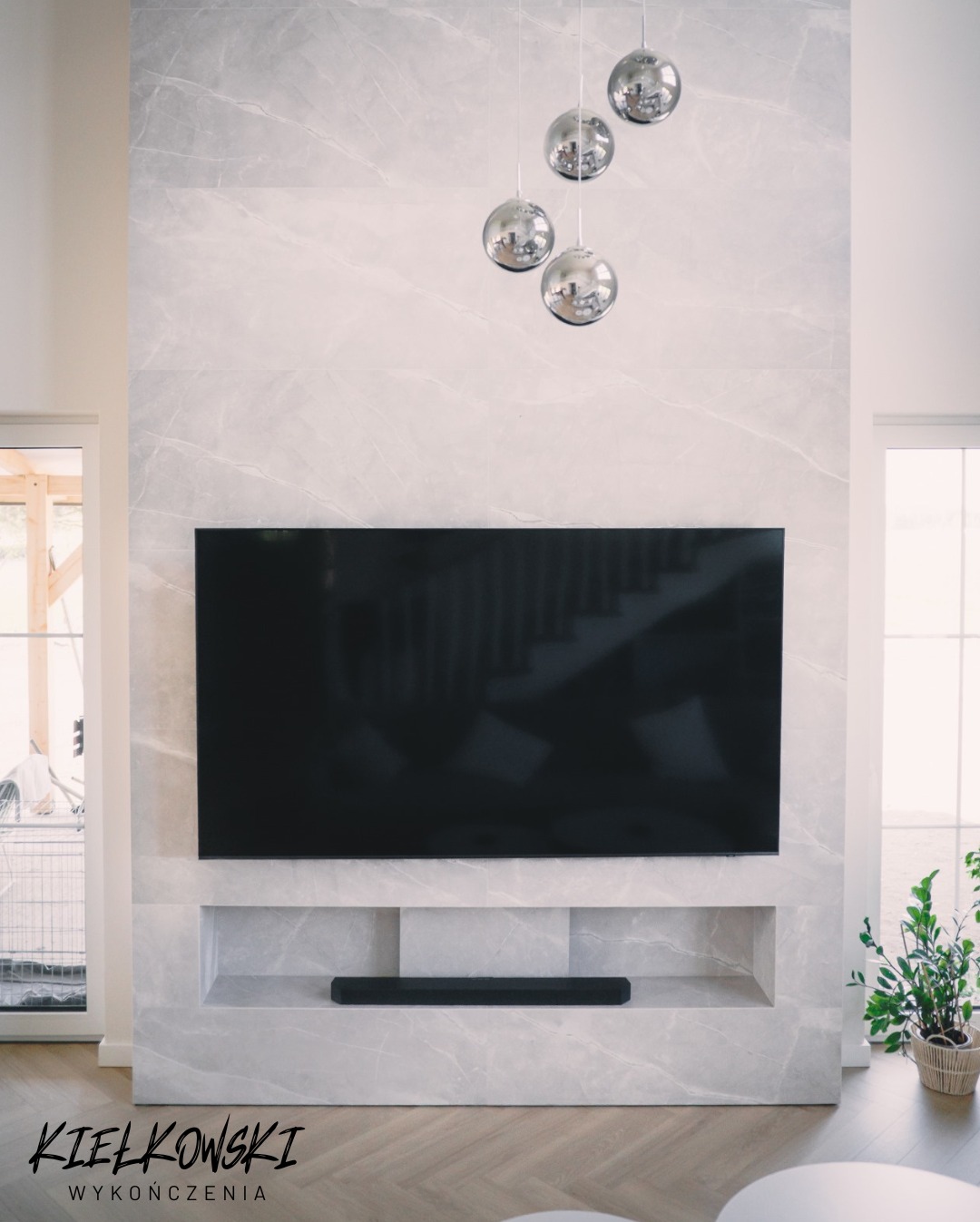Elegancka ściana TV z marmurowym wykończeniem i wnęką na soundbar. Nad telewizorem wiszą cztery chromowane kule-lampy. Parkiet ułożony w jodełkę.