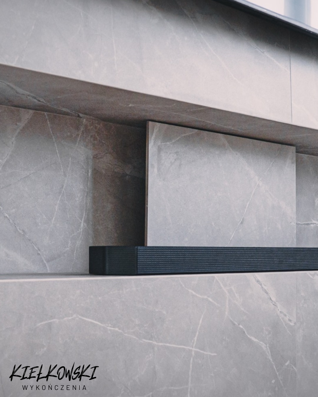 Szara ściana z marmurowym wzorem, wnęka z płytką i czarnym elementem dekoracyjnym. Minimalistyczny design z subtelnym oświetleniem. Kielkowski Wykończenia.
