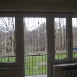 Białe okno PCV z widokiem na balkon z metalową barierką i drzewa w tle, sfotografowane z wnętrza pokoju z widocznym grzejnikiem.