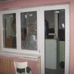 Trzyczęściowe białe okno PCV z naklejkami ochronnymi na szybach, widoczne ślady po montażu na różowej ścianie, grzejnik pod oknem i krzesło w pokoju.