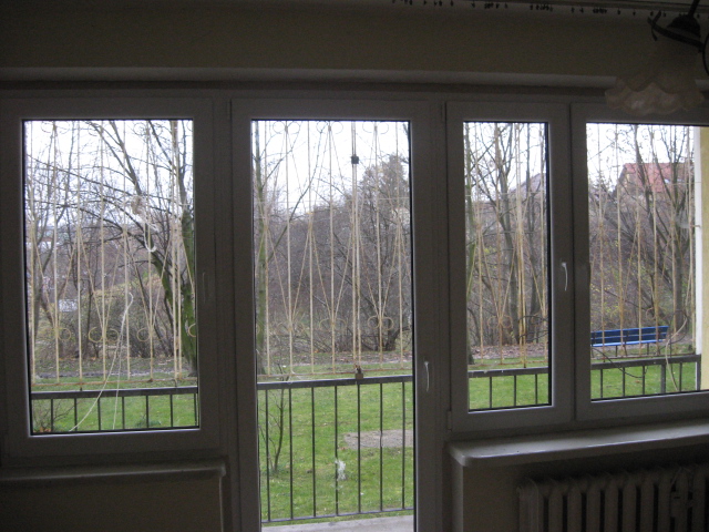 Białe okno PCV z widokiem na balkon z metalową barierką i drzewa w tle, sfotografowane z wnętrza pokoju z widocznym grzejnikiem.