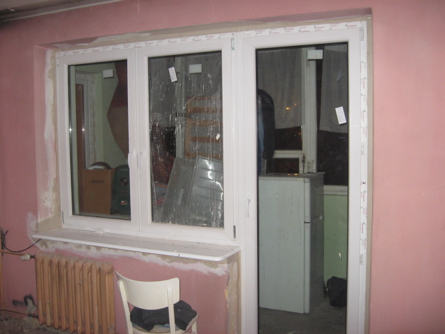 Trzyczęściowe białe okno PCV z naklejkami ochronnymi na szybach, widoczne ślady po montażu na różowej ścianie, grzejnik pod oknem i krzesło w pokoju.