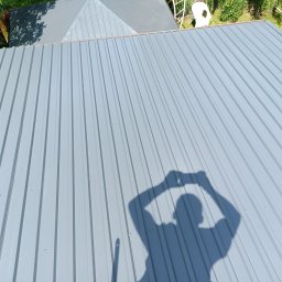 Prace wysokościowe ALPIN - Szary dach z blachy trapezowej, widok z góry, cień osoby robiącej zdjęcie, fragment drabiny i zieleni w tle. Nowe pokrycie dachu.