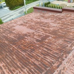 Prace wysokościowe ALPIN - Zardzewiały dach z blachy falistej, widoczne ślady korozji i łuszczącej się farby. Na dachu leży pomarańczowy przewód. W tle zieleń drzew i zabudowa.