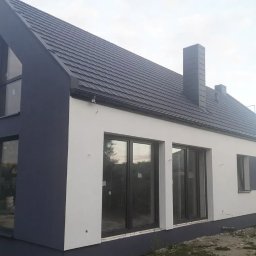 Prace wysokościowe ALPIN - Dwukolorowy dom z ciemnoszarą dachówką i kominem. Biała elewacja kontrastuje z ciemnymi elementami. Widoczne okna i fragment rusztowania.