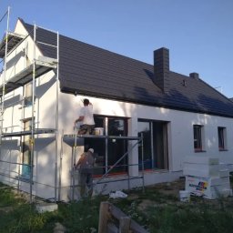 Prace wysokościowe ALPIN - Ekipa maluje elewację domu z rusztowania. Widoczne okna, dach z kominem i materiały budowlane. Jasne tło nieba.