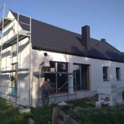 Ekipa maluje elewację domu z rusztowania. Widoczne okna, dach z kominem i materiały budowlane. Jasne tło nieba.