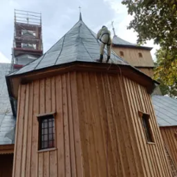 Pracownik w kombinezonie na dachu drewnianego kościoła podczas prac konserwacyjnych. Widoczne rusztowania i drabina oparta o ścianę.