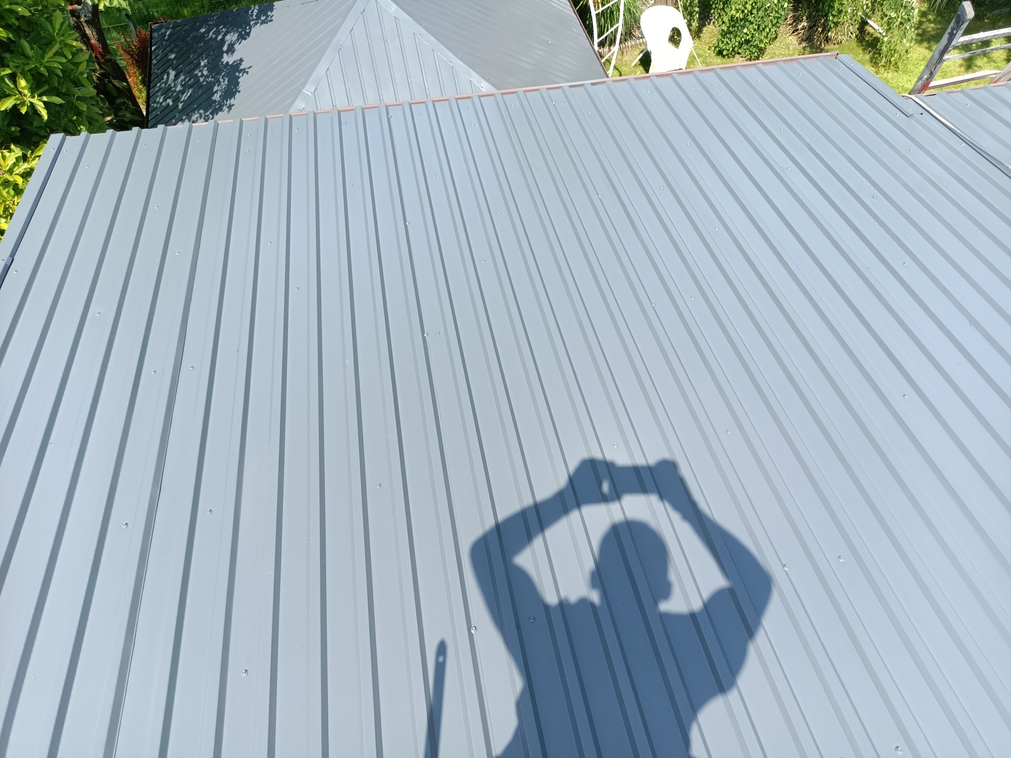Szary dach z blachy trapezowej, widok z góry, cień osoby robiącej zdjęcie, fragment drabiny i zieleni w tle. Nowe pokrycie dachu.