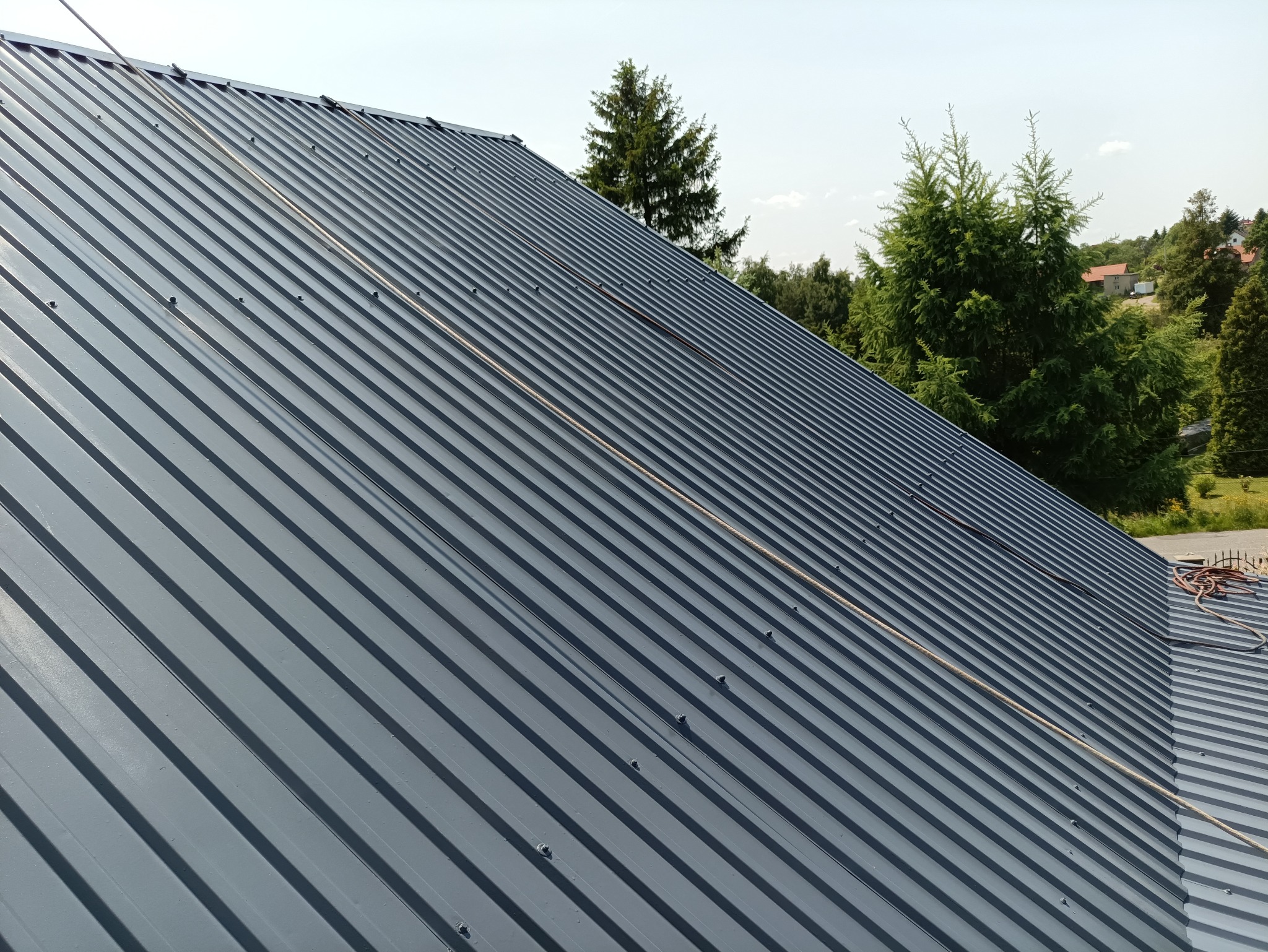 Szary, blaszany dach z widocznymi mocowaniami i linką zabezpieczającą, na tle zieleni drzew. Ujęcie z dołu, podkreślające geometrię dachu.