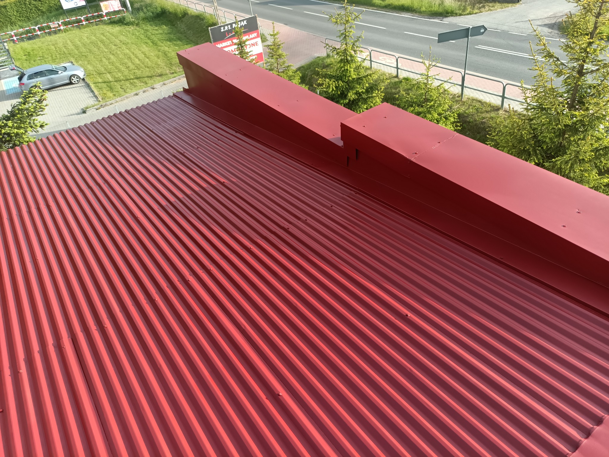 Czerwony, blaszany dach z widocznymi mocowaniami, sfotografowany z góry. W tle zieleń drzew i fragment ulicy.