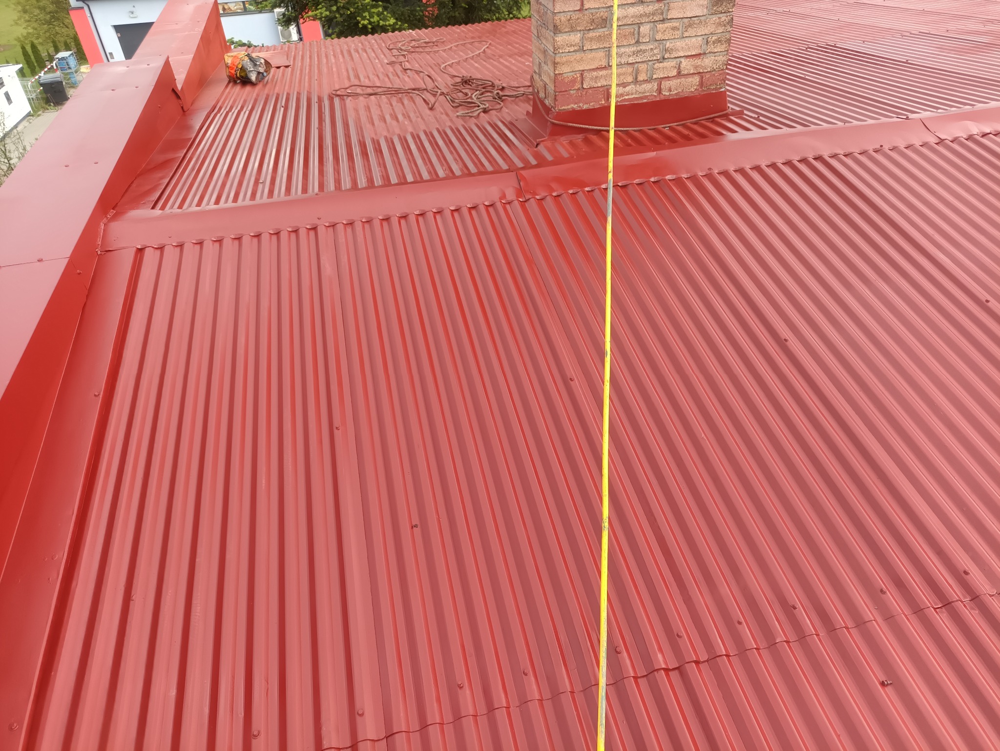 Czerwony, blaszany dach po malowaniu z ceglanym kominem w tle, widziany z góry. Widoczna zwisająca miarka i narzędzia dekarskie. Charakterystyczne karbowanie blachy.