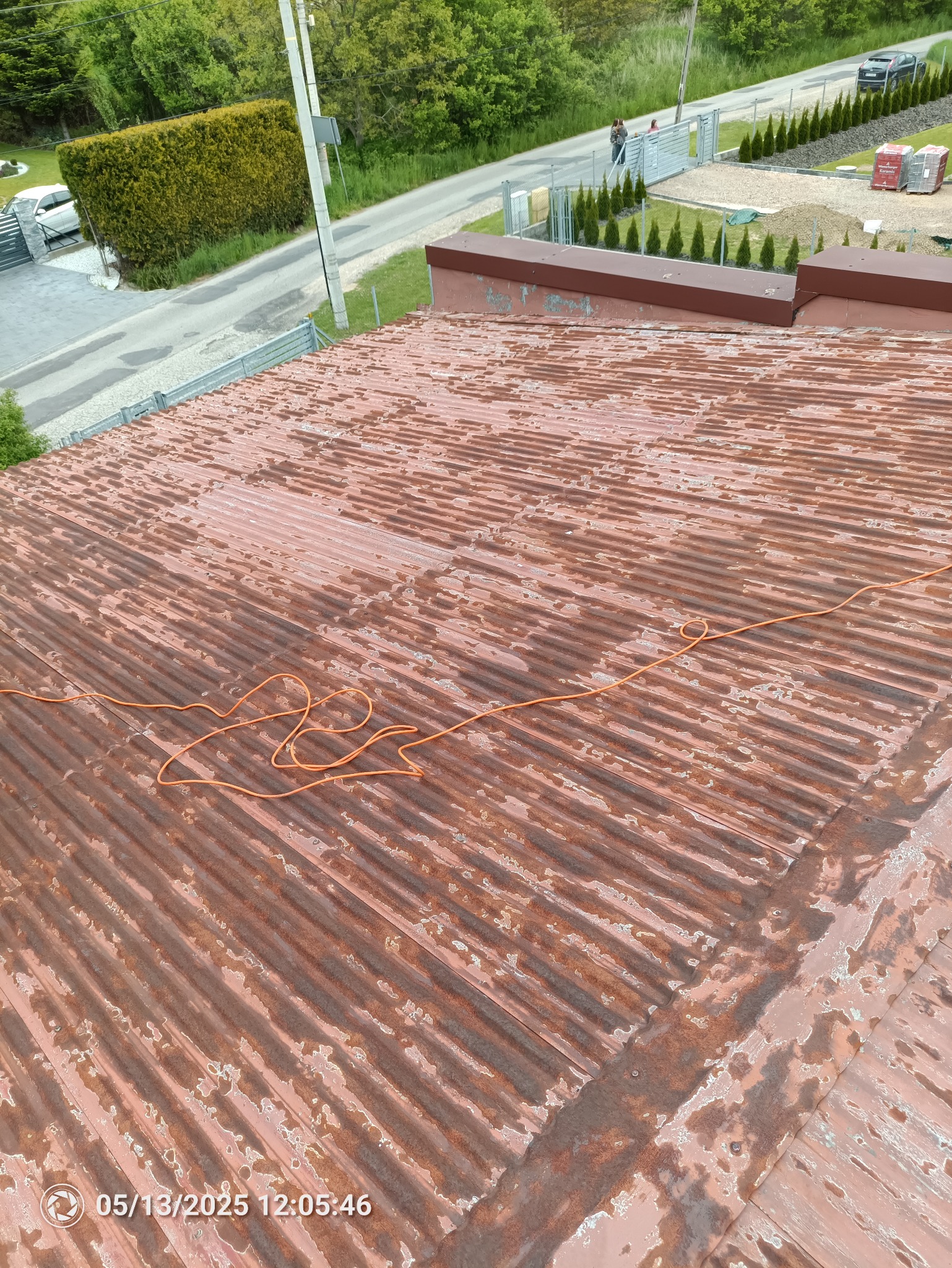 Zardzewiały dach z blachy falistej, widoczne ślady korozji i łuszczącej się farby. Na dachu leży pomarańczowy przewód. W tle zieleń drzew i zabudowa.