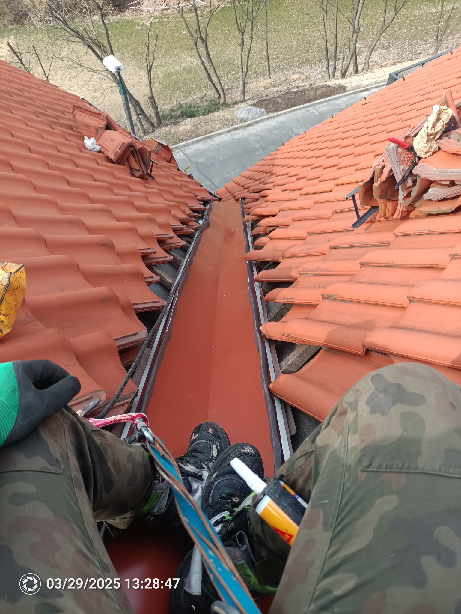 Nowa, pomarańczowa rynna zamontowana między połaciami dachu krytego czerwoną dachówką. Widok z góry na instalatora w uprzęży i spodnie moro.