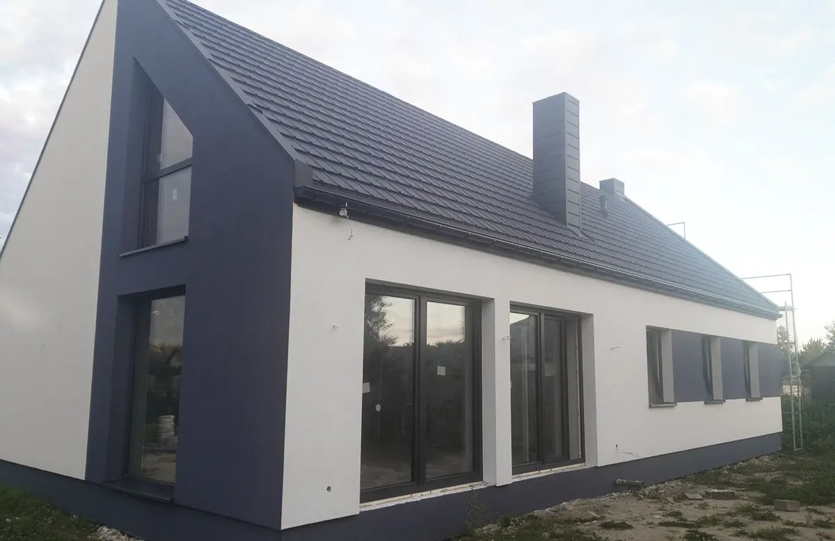 Dwukolorowy dom z ciemnoszarą dachówką i kominem. Biała elewacja kontrastuje z ciemnymi elementami. Widoczne okna i fragment rusztowania.