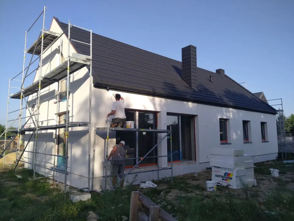 Ekipa maluje elewację domu z rusztowania. Widoczne okna, dach z kominem i materiały budowlane. Jasne tło nieba.