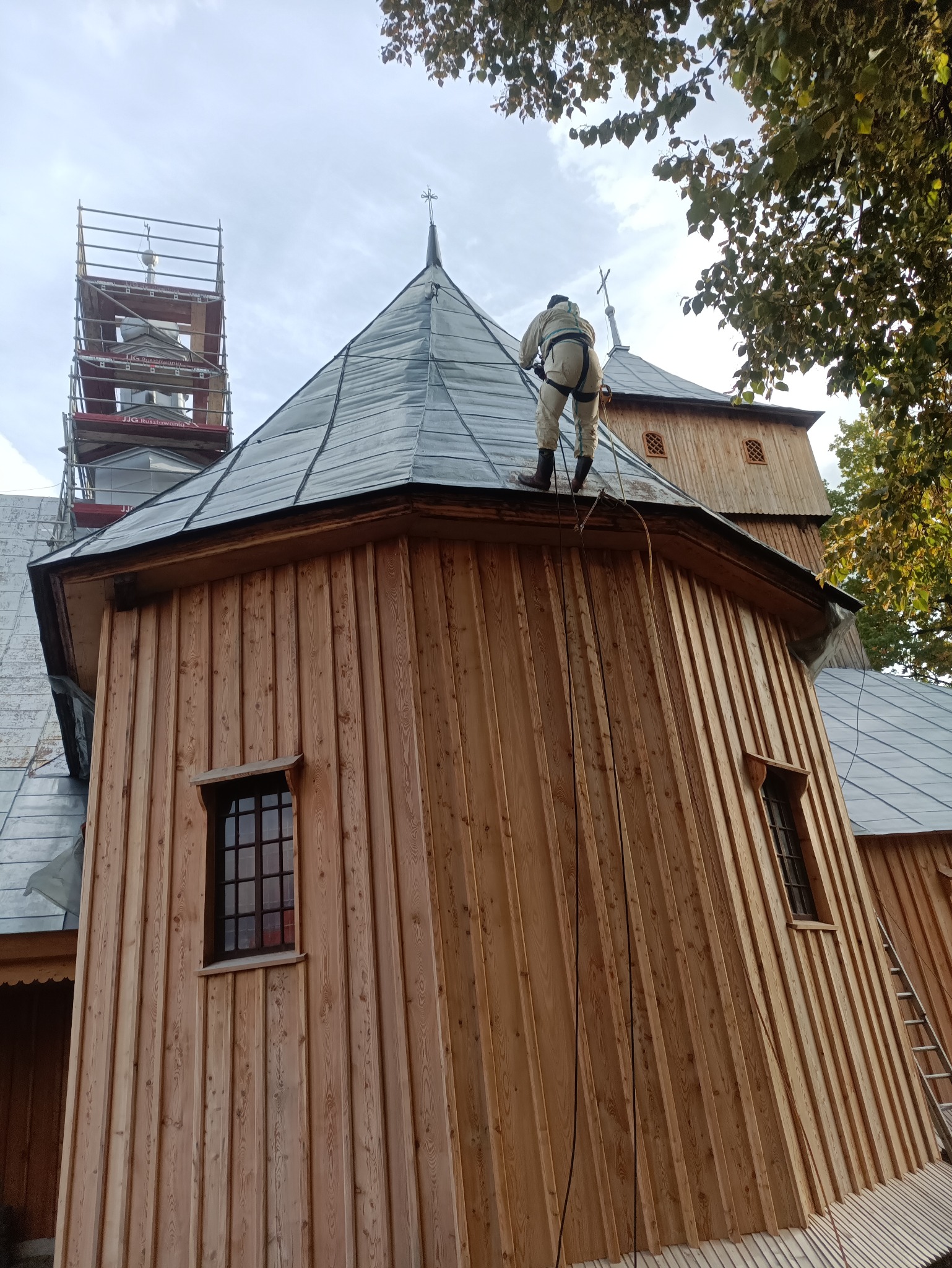 Pracownik w kombinezonie na dachu drewnianego kościoła podczas prac konserwacyjnych. Widoczne rusztowania i drabina oparta o ścianę.