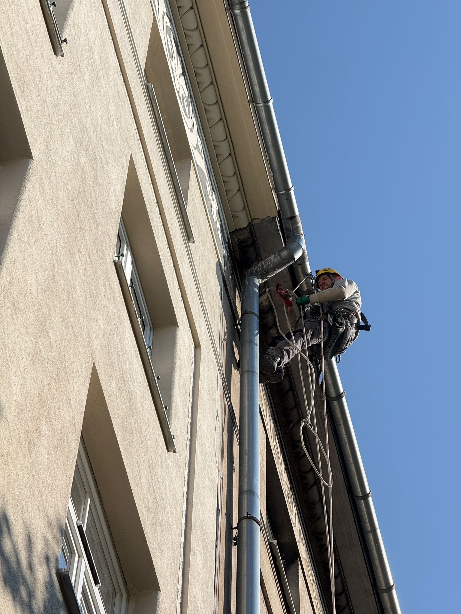 Pracownik w kasku, na linach, czyści rynnę przy elewacji budynku z dekoracyjnymi elementami. Widok z dołu, błękitne niebo w tle. Praca na wysokości.