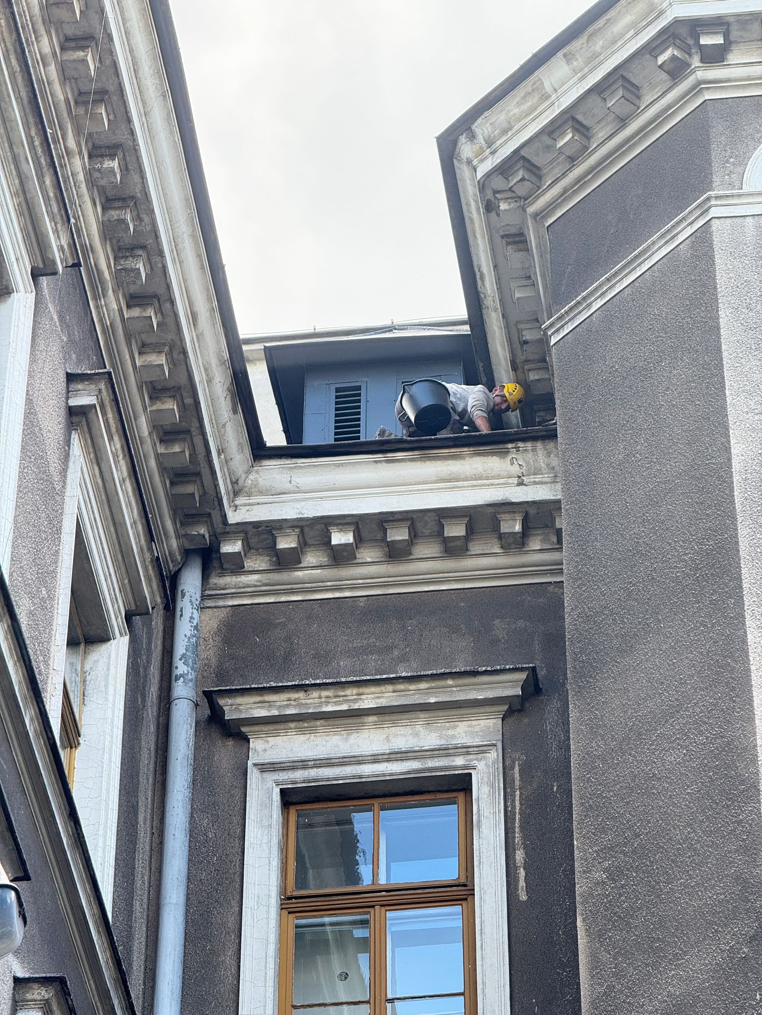 Pracownik w kasku czyści rynnę na zabytkowym budynku z ozdobnymi detalami architektonicznymi, widok z dołu.