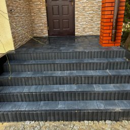 ZIBI BRUK Filip Walas - Schody wejściowe wyłożone ciemną kostką brukową o fakturze prążkowanej, prowadzące do brązowych drzwi wejściowych. Widoczny fragment elewacji z cegły i rynna.