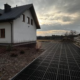 ZIBI BRUK Filip Walas - Metalowa siatka na podłożu obok domu jednorodzinnego, prawdopodobnie wzmocnienie podłoża przed dalszymi pracami. Widoczne przygotowane podłoże i fragment ogrodu.