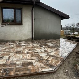 ZIBI BRUK Filip Walas - Ułożona, brązowo-szara kostka na tarasie przy domu. Widoczne elementy wykończeniowe i ziemia wokół. Na dachu kolektory słoneczne. Pochmurne niebo.