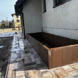 ZIBI BRUK Filip Walas - Ułożona kostka brukowa w odcieniach szarości i brązu, obok brązowa obudowa rabaty kwiatowej przy ścianie domu. Widoczny fragment rynny i trawnik.