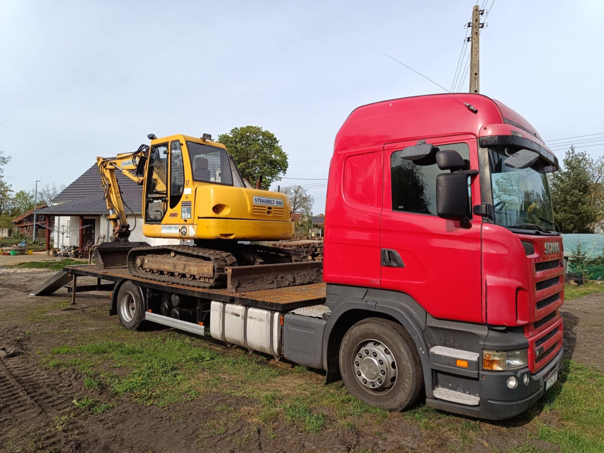 Czerwona ciężarówka Scania z żółtą koparką gąsienicową na platformie transportowej, na tle zielonej trawy i domu.