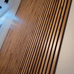 Dekoracyjna ściana z drewnianych listew w ciepłym odcieniu brązu, zamontowana skośnie na białej ścianie, oświetlona przez punktowe lampy sufitowe.